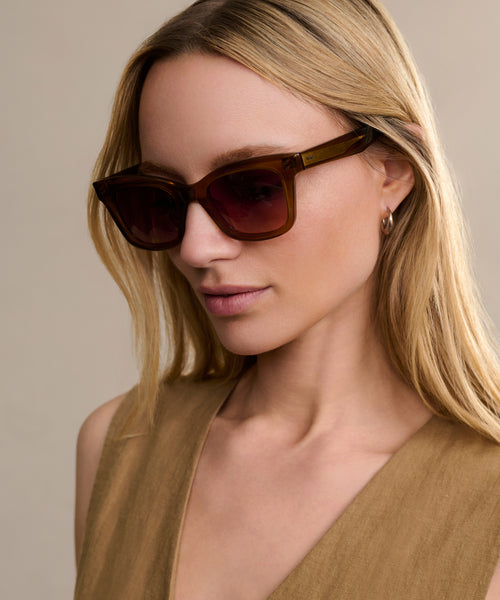 A woman with long blonde hair wears oversized Thompson Sunglasses featuring UV protection, a sleeveless tan top, and small hoop earrings as she poses against a plain beige background.