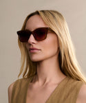 A woman with long blonde hair wears Thompson Sunglasses and a tan sleeveless top, posing against a neutral background.