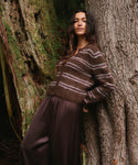 A woman leans against a large tree trunk in a forest setting, wearing a fuzzy brown knit sweater with muted pink and purple horizontal stripes and dark brown satin-like pants.
