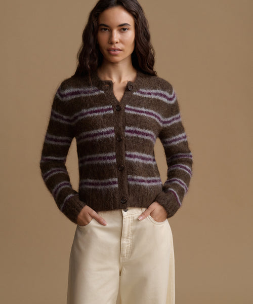 A woman stands against a plain beige studio background wearing a brown knit cardigan with horizontal purple stripes, cream wide-leg pants, and dark brown shoes.