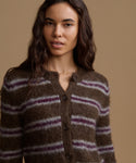 A woman stands against a plain beige studio background wearing a brown knit cardigan with horizontal purple stripes, cream wide-leg pants, and dark brown shoes.