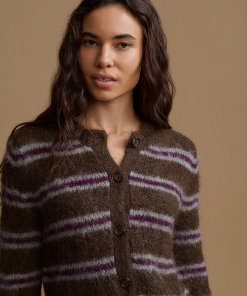 A woman stands against a plain beige studio background wearing a brown knit cardigan with horizontal purple stripes, cream wide-leg pants, and dark brown shoes.