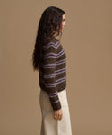 A woman shown in side profile wearing a fuzzy brown knit sweater with horizontal purple and beige stripes, paired with cream-colored pants, against a solid tan studio background.