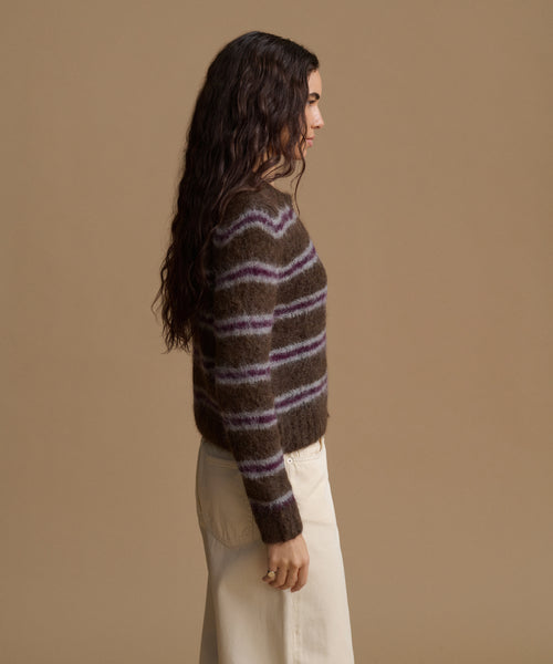 A woman shown in side profile wearing a fuzzy brown knit sweater with horizontal purple and beige stripes, paired with cream-colored pants, against a solid tan studio background.