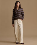 A woman stands against a plain beige studio background wearing a brown knit cardigan with horizontal purple stripes, cream wide-leg pants, and dark brown shoes.