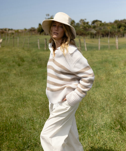 A woman wearing the Cashmere Phoebe Polo and white pants stands in a grassy field with a fence and trees in the background.