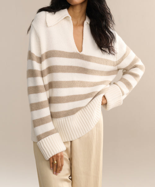 A person models the Cashmere Phoebe Polo—an ivory striped collared sweater—paired with beige pants, standing with one hand on their hip against a neutral backdrop.