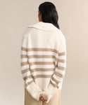 A person with long dark hair faces away, wearing the Cashmere Phoebe Polo in ivory stripe with a large ribbed collar. Their hands are clasped behind their back against a neutral beige background.