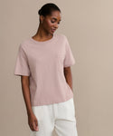 Wearing the Vintage Pocket Tee in light pink with a chest pocket and white pants, this look against a beige background and subtle smile makes it an ideal everyday wardrobe choice.