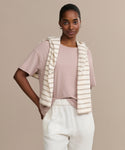 A woman wears the Vintage Pocket Tee in light pink, paired with white pants and a cream-beige striped sweater over her shoulders, standing against a beige background and smiling softly with one hand in her pocket.