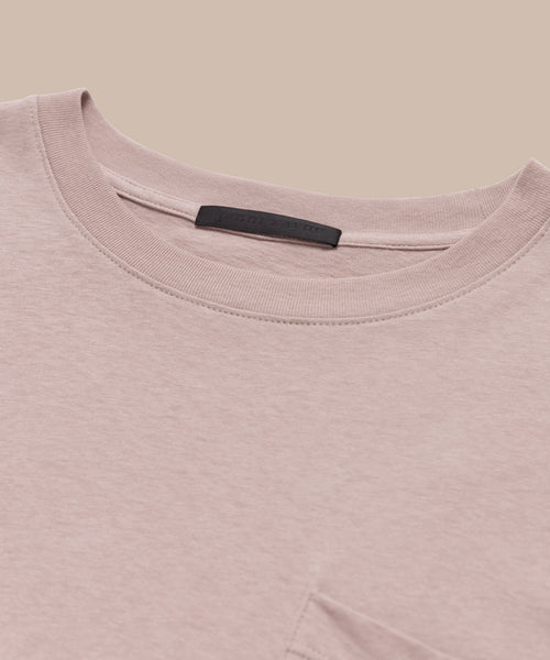 Close-up of the Vintage Pocket Tee in light pink, featuring a chest pocket and black collar label, laid flat on a beige background—ideal for everyday wear.