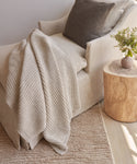 The Wool Fisherman Throw in beige, crafted from soft Andean Highland wool in Peru, is draped over a cream armchair with a brown cushion, beside a wooden table holding a vase of greenery on a light rug and wooden floor.