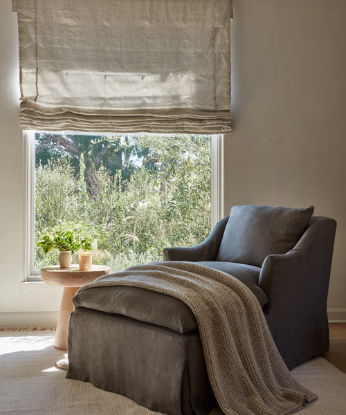 A cozy armchair with a matching ottoman and a Wool Fisherman Throw sits by a sunlit window. Nearby, a wooden side table holds plants and cups while light streams through a linen Roman shade, revealing greenery outside.