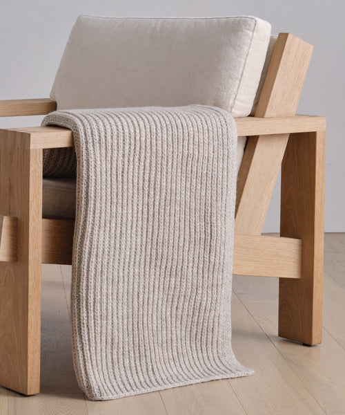 A modern wooden chair with cream cushions features the Wool Fisherman Throw draped over one arm, set on a light wood floor against a plain wall.