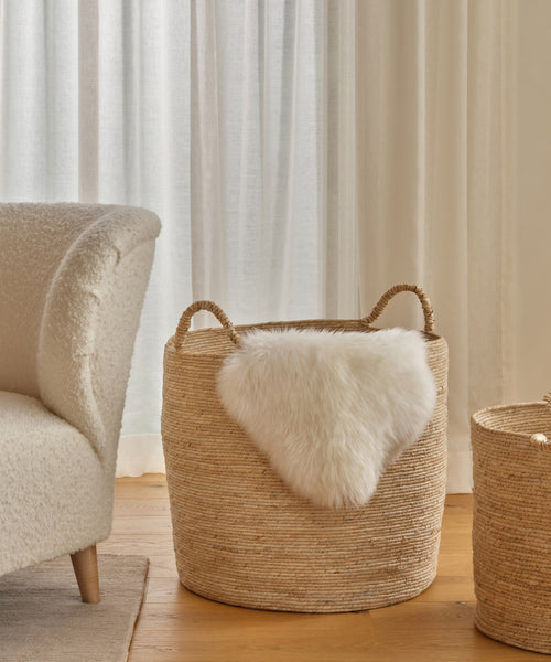 The Woven Storage Basket with rope handles sits on a wooden floor beside a cream armchair. A white faux fur throw drapes over it, while sheer curtains let in soft light, making this basket the perfect home storage accent.