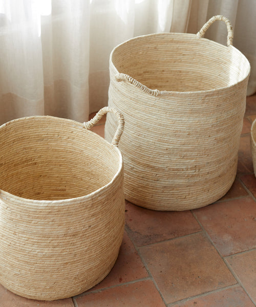 Two large Woven Storage Baskets with handles, in a natural color, rest on terracotta tiles beside sheer white curtains, providing stylish home storage.