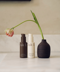 Two bottles of the Youthful Skin Serum Duo stand on a marble surface, promoting skin renewal and hydration. They are beside a small dark vase with a single drooping pink flower, set against a neutral background.
