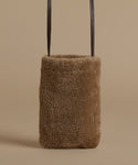 A small rectangular shoulder bag made of soft brown shearling, with dark brown leather shoulder straps, shown against a solid beige background.