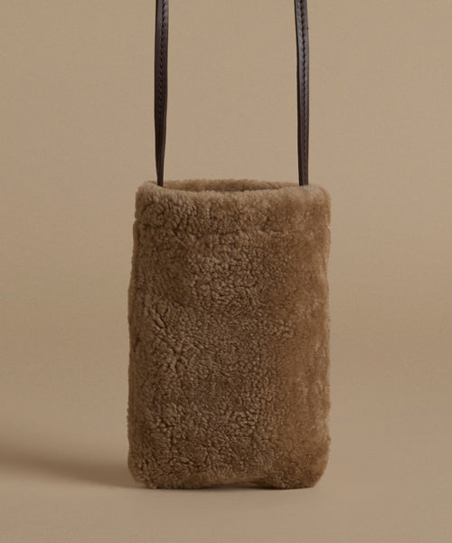 A small rectangular shoulder bag made of soft brown shearling, with dark brown leather shoulder straps, shown against a solid beige background.