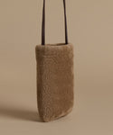 A vertical, rectangular shoulder bag made of soft, textured shearling in a warm taupe-brown color, featuring two slim dark brown leather straps, set against a solid beige background.
