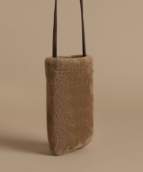A vertical, rectangular shoulder bag made of soft, textured shearling in a warm taupe-brown color, featuring two slim dark brown leather straps, set against a solid beige background.