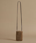 A small cylindrical shoulder bag made of brown textured shearling with a thin black shoulder strap, set against a solid beige background.