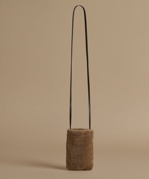 A small cylindrical shoulder bag made of brown textured shearling with a thin black shoulder strap, set against a solid beige background.