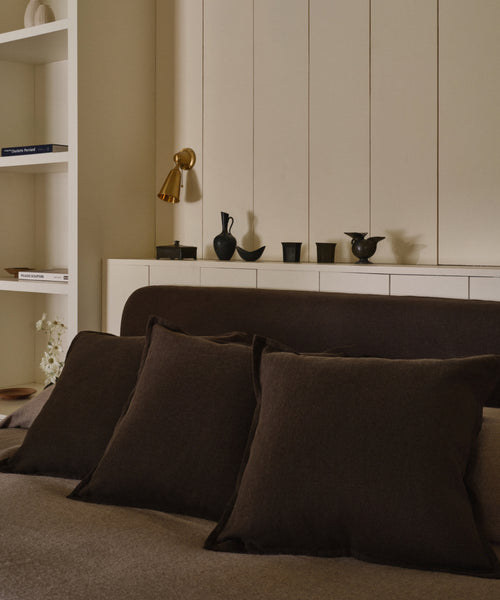 A cozy bedroom with a bed featuring three Adler Pillows in dark brown Belgian linen, a beige headboard, a gold wall lamp, minimalist black decor on a white shelf, and books on a built-in bookshelf.