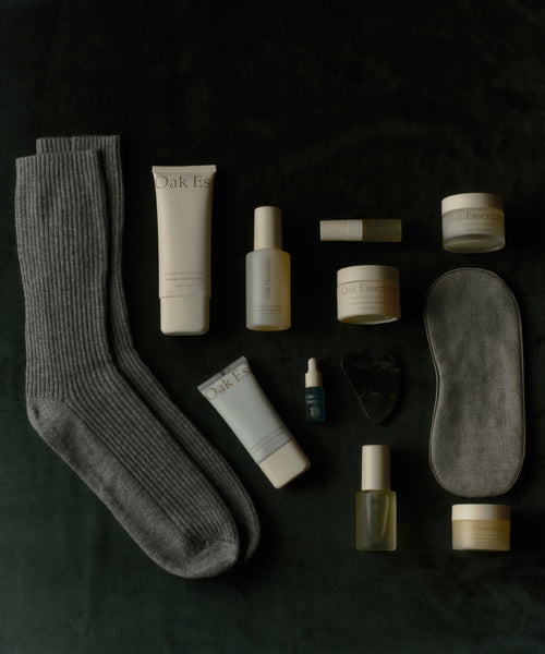 A flat lay of wellness items on a dark surface, featuring the Oak Essentials x Jenni Kayne Advent Calendar—a curated set with grey ribbed socks, beige and gold skincare, a blue serum bottle, gua sha tool, sleep mask, and lip balm for perfect self-care gifting.