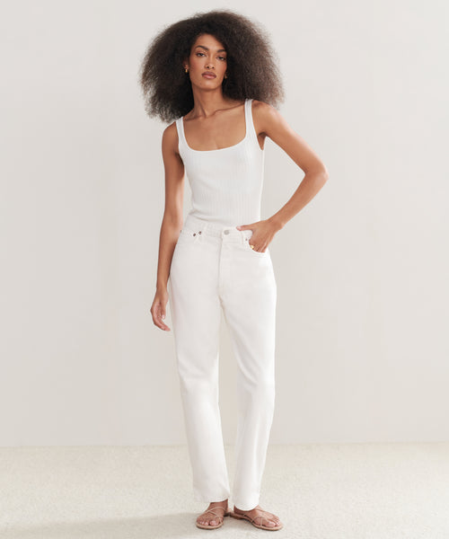 A woman with curly hair wears a white tank top, AGOLDE 90s Jean in organic cotton denim, and brown sandals, standing against a plain light background.