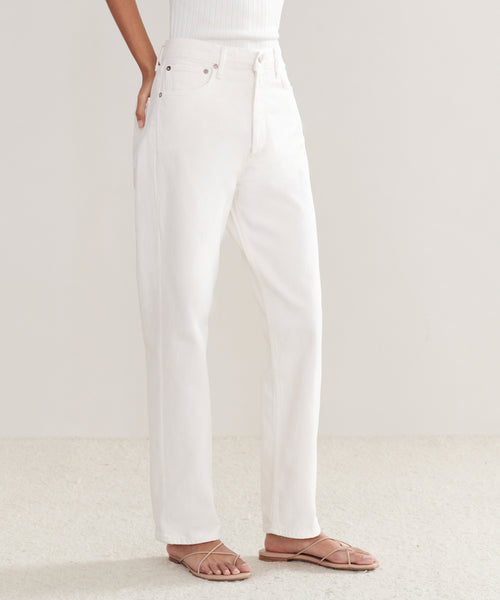 Wearing the 90s Jean—vintage-inspired, high-waisted straight-leg white jeans—a person pairs them with a white ribbed tank and strappy sandals in a minimal, light-toned room, their hand casually resting in the pocket.