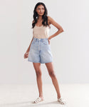 A woman with long dark hair and a neutral expression stands indoors against a plain light background, wearing the Stella Short—a pair of vintage denim high-waisted shorts—paired with a beige tank top and white flat sandals, hand on her hip.