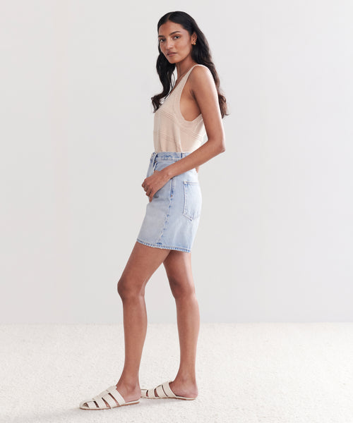 A woman with long, wavy dark hair stands in profile against a light background, wearing the Stella Short in beige, paired with a vintage denim skirt and white slide sandals.
