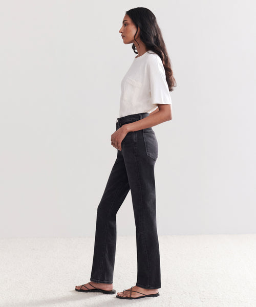 A woman in profile against a plain background wears a white short-sleeve T-shirt, High Rise Stovepipe Jean, and black sandals. She has long dark hair and her hands are in her pockets.