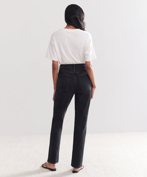 A person with long dark hair faces away, wearing a white short-sleeve t-shirt, High Rise Stovepipe Jean, and flat sandals, set against a plain light background and carpeted floor.