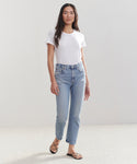A woman with long, straight hair and a relaxed expression stands against a plain light background wearing a white t-shirt, black strappy sandals, and the High Rise Stovepipe Jean in vintage denim.
