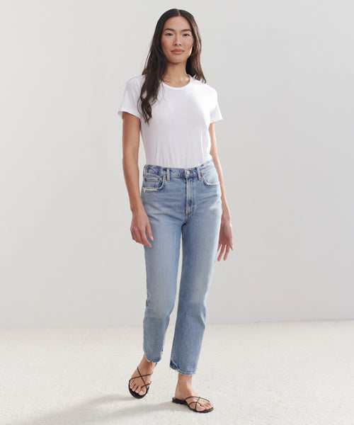 A woman with long, straight hair and a relaxed expression stands against a plain light background wearing a white t-shirt, black strappy sandals, and the High Rise Stovepipe Jean in vintage denim.