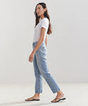 A woman in profile stands against a light background wearing a white t-shirt, the High Rise Stovepipe Jean in vintage denim, and black sandals. Her loose long hair complements her neutral expression.