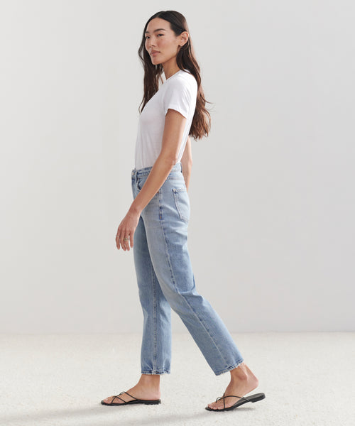 A woman in profile stands against a light background wearing a white t-shirt, the High Rise Stovepipe Jean in vintage denim, and black sandals. Her loose long hair complements her neutral expression.