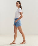 A woman stands sideways, smiling, in a white t-shirt, Parker Long Short by AGOLDE denim vintage fit shorts, and brown sandals. She poses indoors against a plain light-colored background.