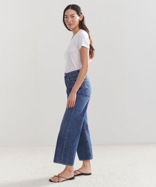 A woman with long dark hair stands sideways in a white tee, black sandals, and the high-waisted vintage denim Ren Jean, set against a plain light background.