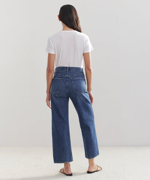 A woman with long dark hair, dressed in a white t-shirt and Ren Jeans, stands facing away from the camera against a light gray background, completing her look with classic black sandals.