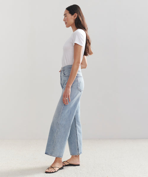 A woman stands in profile against a plain background, wearing a white short-sleeve t-shirt, black sandals, and high-waisted AGOLDE Ren Jean denim jeans in light blue.