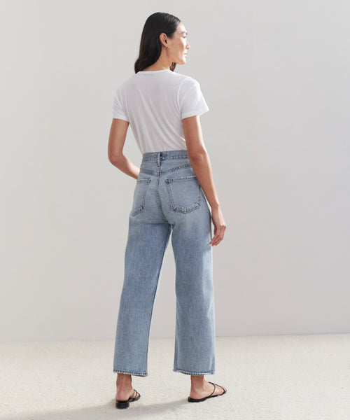 A person stands facing away, wearing a white t-shirt, black sandals, and the Ren Jean—high-waisted vintage jeans. The background is plain and light-colored.