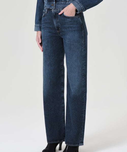 A person stands against a plain white background wearing Harper Straight Jean in relaxed mid-rise, dark blue, straight-leg style with black heeled shoes and one hand in their pocket.