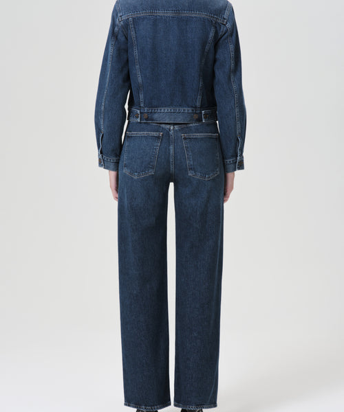 A person stands facing away, wearing the Harper Straight Jean in dark blue denim with visible seams and pockets, paired with a matching jacket against a plain light background.