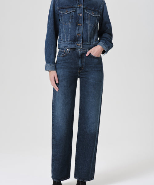A person stands with one hand in their pocket, wearing dark blue matching organic cotton denim and the Harper Straight Jean, featuring a high waist and wide-leg fit, against a plain light background.