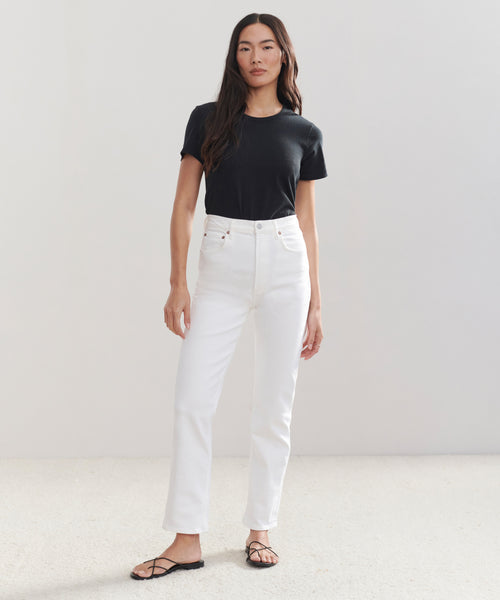 A woman with long dark hair stands against a light background, wearing a black short-sleeve t-shirt, High Rise Stovepipe Jean in vintage denim, and black sandals. She has a neutral expression.