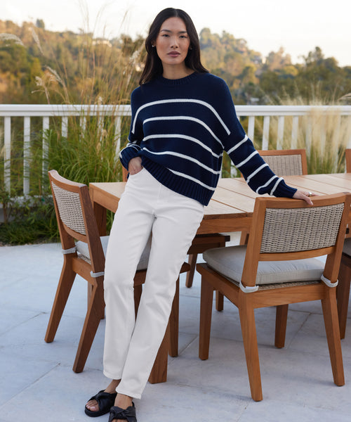 Outdoors by a wooden patio set, a woman in a navy blue sweater with white stripes and High Rise Stovepipe Jean looks calmly at the camera, surrounded by greenery and trees.