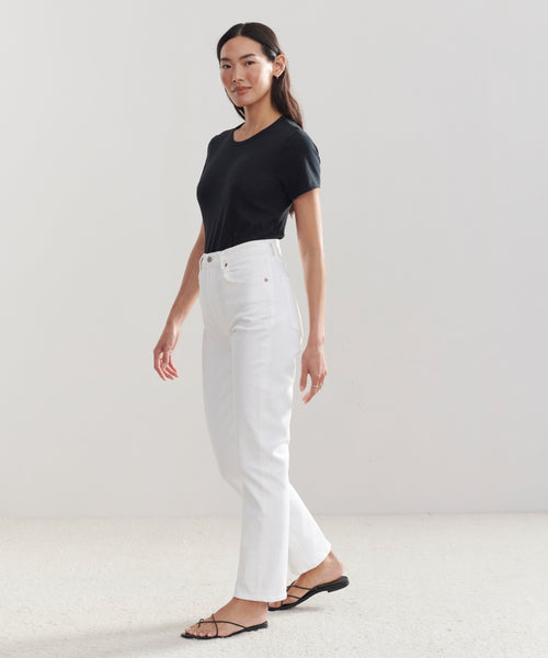 A woman with long dark hair wears a black t-shirt, High Rise Stovepipe Jean in vintage denim, and black sandals against a light background, looking to the side with a relaxed expression.